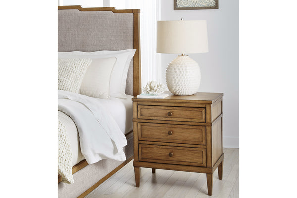 Wooden nightstand of three drawers with decorative objects on top with a bed to the left in a bright bedroom setting.