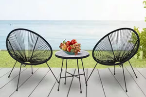 Two black woven chairs with a small round table on a wooden deck with a scenic background.