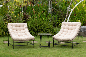 Two white cushioned outdoor chairs with rattan bodies and with a small table with glass top on a grassy area with greenery in the background.