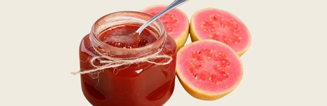 Envase con salsa de guayaba adentro con cuchara y tres guayabas por el lado derecho, todo sobre un fondo crema.