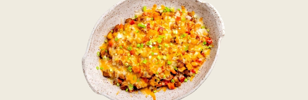Cazuela de papas con queso rallado y tocineta encima servido en un plato grande crema, todo sobre un fondo crema.