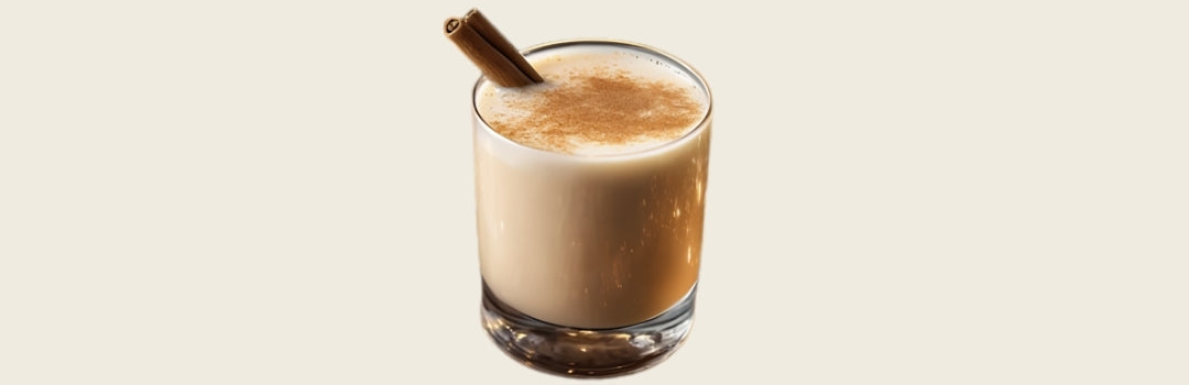 Coquito un rama de canela en el borde adentro de un vaso, todo sobre un fondo crema.