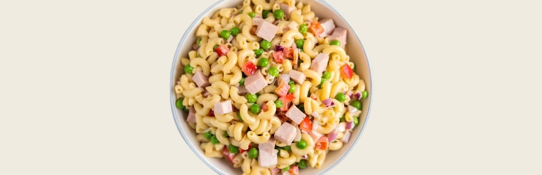 Ensalada de coditos con cantos de jamón, pimientos y guisantes sobre un fondo crema. 
