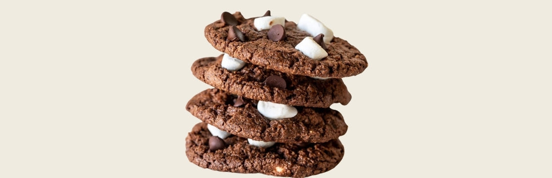 Galletas de chocolate con marshmallows y chispas de chocolate encima de otros, todo sobre un fondo blanco.