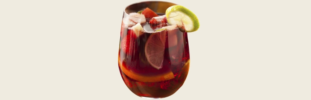 Sangría con fresas y pedazos de piñas adentro con una rodaja de limón en el borde, todo sobre un fondo crema.