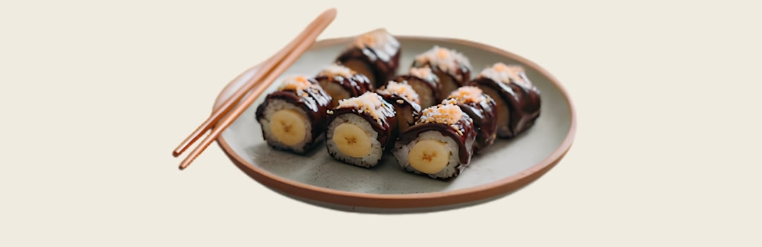 Varios pedazos de sushi con guineo adentro y cubierto en chocolate servido en un plato con palillos, todo sobre un fondo crema. 
