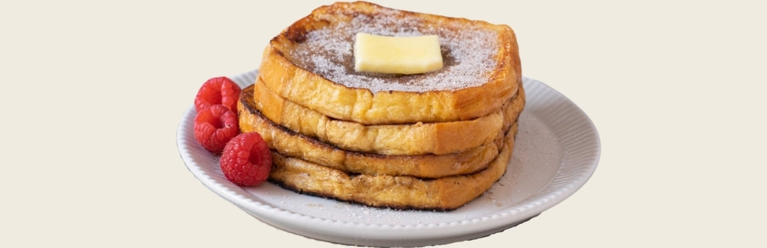 Cuatro tostadas francesas con mantequilla y syrup encima y con frambuesas por el lado servido en un plato blanco sobre un fondo crema.