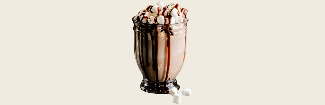 Coquito de chocolate con marshmallows por encima serviddo en un vaso de vidro, todo sobre un fondo crema.