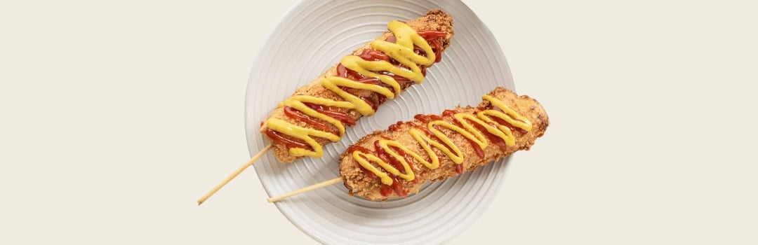 Dos hot dog coreanos con ketchup y mostaza encima servido en un plato, todo sobre un fondo crema.