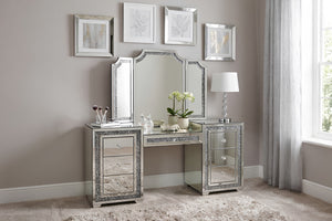 Vanity glamuroso con acabado de espejo y detalles brillantes, acompañado de espejo tríptico y cajones decorativos. Ambientado en un dormitorio elegante con lámpara de mesa, cuadros enmarcados y alfombra color crema.
