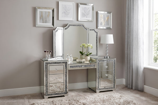 Vanity glamuroso con acabado de espejo y detalles brillantes, acompañado de espejo tríptico y cajones decorativos. Ambientado en un dormitorio elegante con lámpara de mesa, cuadros enmarcados y alfombra color crema.