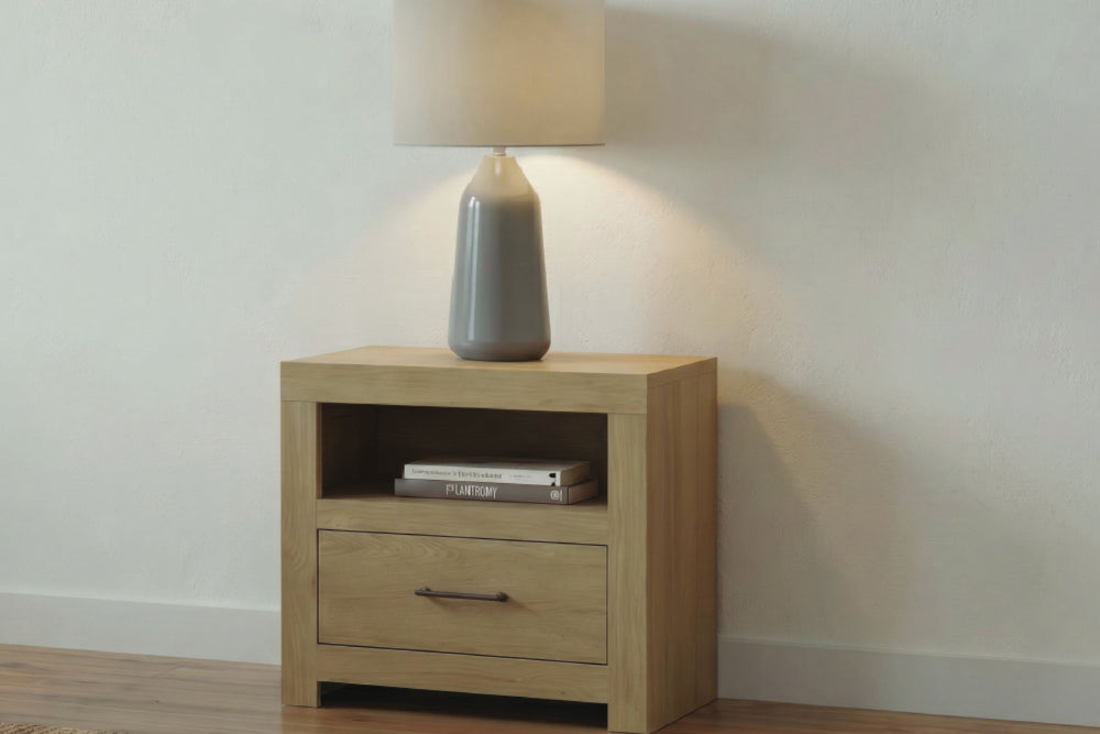 Mesa de noche clara con una gaveta y estante abierto, decorada con una lámpara de mesa de cerámica gris y libros, ubicada frente a una pared neutra color blanco roto y un suelo de madera clara.