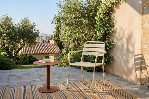 Una silla de comedor para patio de metal beige claro está colocada sobre una alfombra tejida junto a una pequeña mesa auxiliar redonda de madera con una bebida, situada en un patio de piedra con olivos y una pared de estuco al fondo.