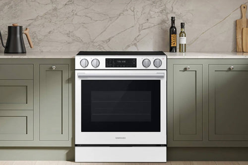 Una estufa eléctrica Samsung blanca con cubierta y puerta de horno, ubicada entre gabinetes de cocina verde salvia contra un counter de mármol.