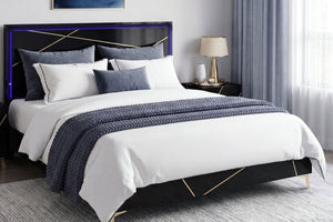 Una cama moderna con estructura negra y detalles geométricos dorados, vestida con ropa de cama blanca y una manta decorativa azul marino texturizada, ubicada en un dormitorio decorado con estilo.