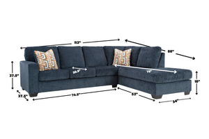 Sofá seccional en forma de L tapizado en azul oscuro con chaise longue a la derecha y cojines decorativos geométricos. La imagen incluye medidas del mueble, como ancho, profundidad, altura y dimensiones de los asientos, sobre fondo neutro.