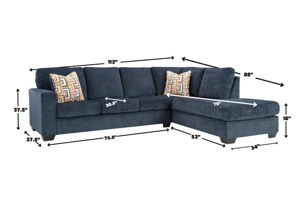 Sofá seccional en forma de L tapizado en azul oscuro con chaise longue a la derecha y cojines decorativos geométricos. La imagen incluye medidas del mueble, como ancho, profundidad, altura y dimensiones de los asientos, sobre fondo neutro.