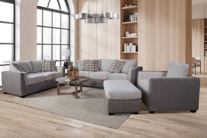 Juego de sala modular en tela gris con sofá, loveseat, silla de acento y otomana, acompañado de cojines decorativos, presentado en una sala moderna con ventanales amplios.