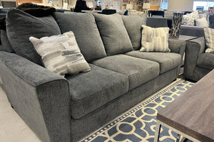 Sofá de tres plazas tapizado en tela gris oscuro con cojines de respaldo amplios y cojines decorativos estampados en tonos grises. La imagen destaca los asientos acolchados y el diseño cómodo del mueble en una exhibición de tienda.