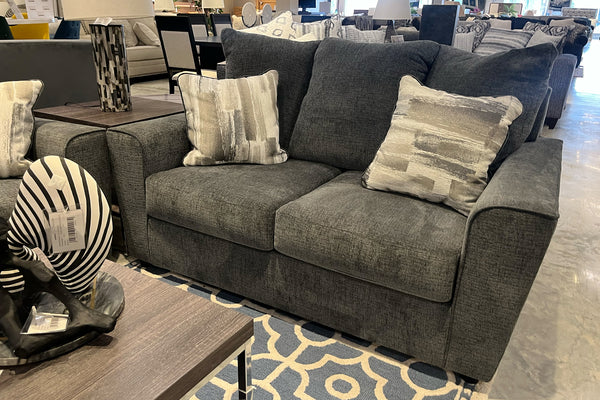 Loveseat tapizado en tela gris oscuro con cojines de respaldo amplios y cojines decorativos estampados en tonos grises. El mueble se exhibe sobre una alfombra geométrica azul y beige junto a una mesa de centro en una tienda de muebles.