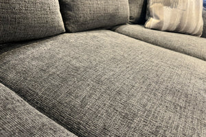 Primer plano del asiento y respaldo de un sofá tapizado en tela gris texturizada. La imagen resalta el tejido suave, los cojines acolchados y la comodidad del mueble, con un cojín decorativo visible en uno de los extremos.