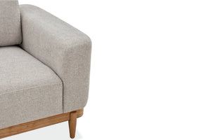 Primer plano de la esquina de un sillón tapizado en color gris claro, con pata de madera natural sobre un fondo blanco.