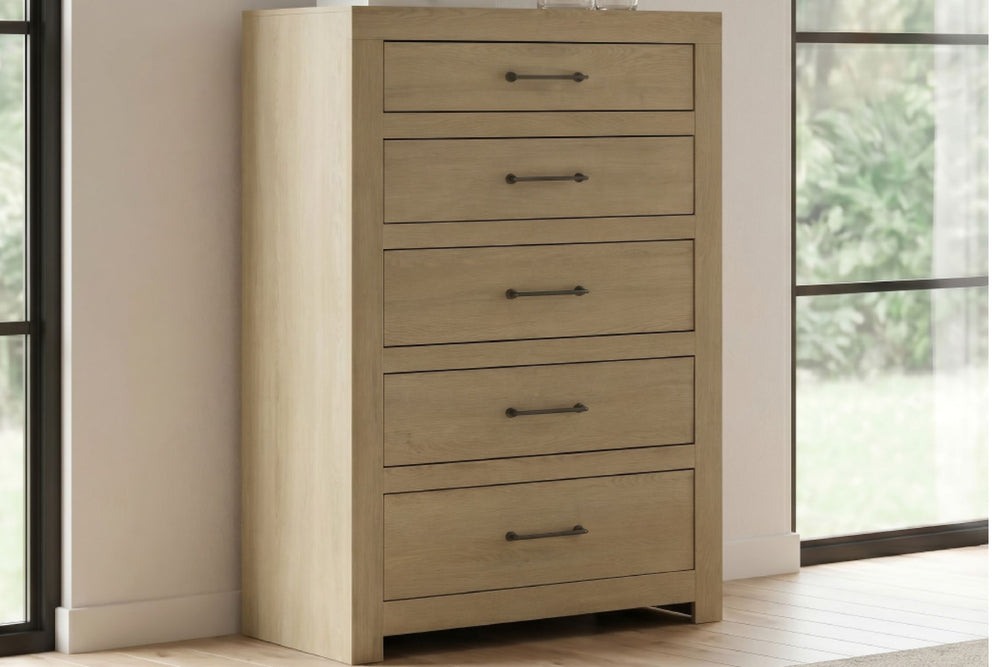 Chest de madera clara con 5 gavetas y agarraderas de barra de metal oscuro, ubicada en la ambientación de un dormitorio entre dos ventanas.