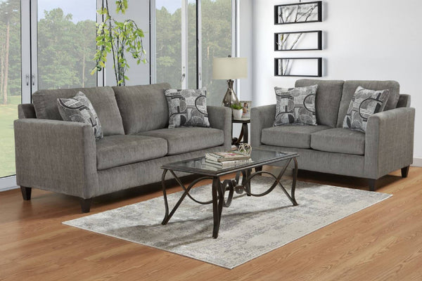 Juego de sala moderno en color gris con sofá de tres plazas y loveseat de dos plazas, ambos con cojines decorativos estampados y apoyabrazos rectos, ubicado en una sala luminosa con mesa de centro de vidrio y ventanales amplios.