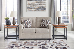 Loveseat de color beige con cojines decorativos y con elementos decorativos en una sala con ventanas en el fondo. 