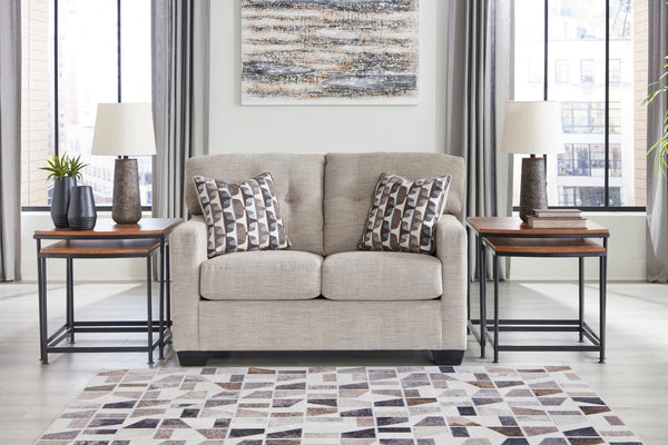 Loveseat de color beige con cojines decorativos y con elementos decorativos en una sala con ventanas en el fondo. 