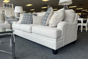 Sofá moderno tapizado en tela beige clara con cojines decorativos en patrones florales y cuadros en tonos gris, azul y crema, acompañado de lámparas de mesa y muebles de exhibición en una tienda.
