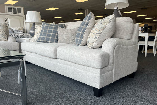Sofá moderno tapizado en tela beige clara con cojines decorativos en patrones florales y cuadros en tonos gris, azul y crema, acompañado de lámparas de mesa y muebles de exhibición en una tienda.
