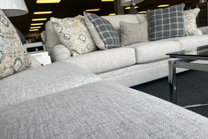 Sala de exhibición con sofá y loveseat tapizados en tela beige clara, cojines decorativos con patrones florales y cuadros en tonos gris, azul y crema, y mesa de centro con cubierta de vidrio y estructura metálica.