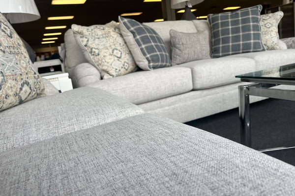 Sala de exhibición con sofá y loveseat tapizados en tela beige clara, cojines decorativos con patrones florales y cuadros en tonos gris, azul y crema, y mesa de centro con cubierta de vidrio y estructura metálica.