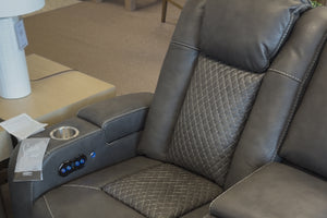 Primer plano de loveseat reclinable tapizado en cuero sintético gris oscuro, con respaldo acolchado de patrón romboidal, portavasos metálico integrado y panel lateral con controles iluminados para ajuste eléctrico.