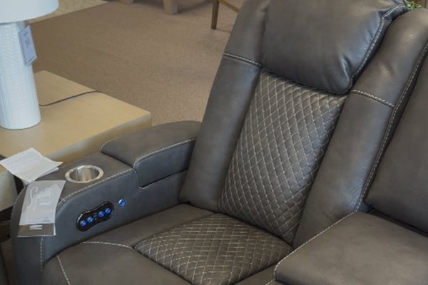 Primer plano de loveseat reclinable tapizado en cuero sintético gris oscuro, con respaldo acolchado de patrón romboidal, portavasos metálico integrado y panel lateral con controles iluminados para ajuste eléctrico.