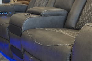 Primer plano de sofá reclinable tapizado en cuero sintético gris oscuro, con asiento acolchado, costuras decorativas en contraste, portavasos integrado e iluminación LED azul en la base.