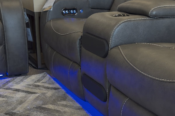 Primer plano de sofá reclinable tapizado en cuero sintético gris oscuro, con asiento acolchado, costuras decorativas en contraste, portavasos integrado e iluminación LED azul en la base.