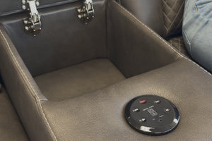 Primer plano de consola central de sofá reclinable tapizado en cuero sintético gris oscuro, con compartimiento de almacenamiento abierto y panel de controles táctiles integrados sobre el apoyabrazos.
