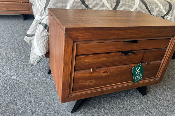 Mesa de noche de madera en tono marrón medio con acabado visible de vetas, diseño rectangular y dos gavetas frontales con tiradores horizontales oscuros. Incluye un detalle decorativo en el cajón inferior y una etiqueta colgante con certificación FSC. La pieza se muestra junto a una cama con ropa de cama en tonos neutros, sobre alfombra gris en un entorno de exhibición.