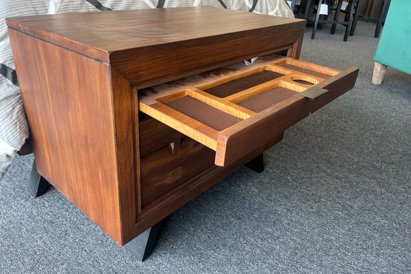 Mesa de noche de madera en tono marrón con dos gavetas; la gaveta superior está abierto y muestra un organizador interior con compartimentos para objetos pequeños. La gaveta presenta vetas visibles, tiradores horizontales oscuros y patas inclinadas, ubicado junto a una cama sobre superficie de alfombra en un entorno de exhibición.
