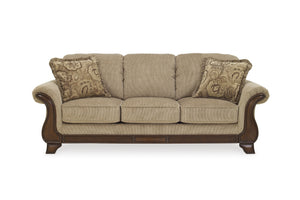 Un sofá de tres plazas con tapicería texturizada de color beige, brazos y patas de madera marrón oscuro y dos cojines decorativos con estampado de cachemir, mostrados sobre un fondo blanco liso.