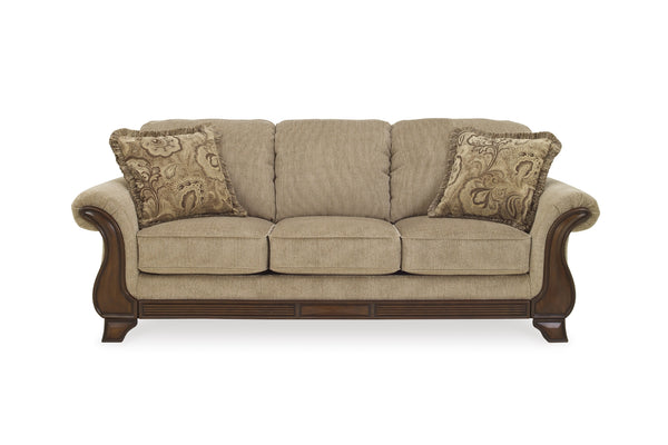 Un sofá de tres plazas con tapicería texturizada de color beige, brazos y patas de madera marrón oscuro y dos cojines decorativos con estampado de cachemir, mostrados sobre un fondo blanco liso.