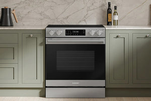Una estufa eléctrica Samsung con una placa de cocina de vidrio negro y puerta de horno, ubicada entre gabinetes verde salvia en una cocina moderna.