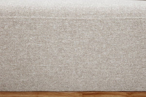 Primer plano de un tapizado de tela texturizada en beige claro, con patrón tejido y una costura fina. En la parte inferior se observa una franja de piso de madera marrón cálido.