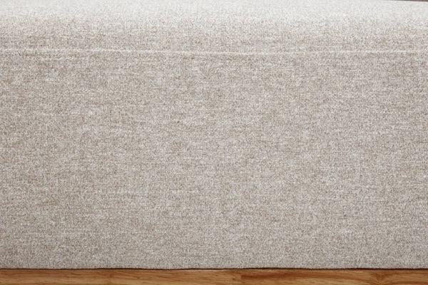 Primer plano de un tapizado de tela texturizada en beige claro, con patrón tejido y una costura fina. En la parte inferior se observa una franja de piso de madera marrón cálido.