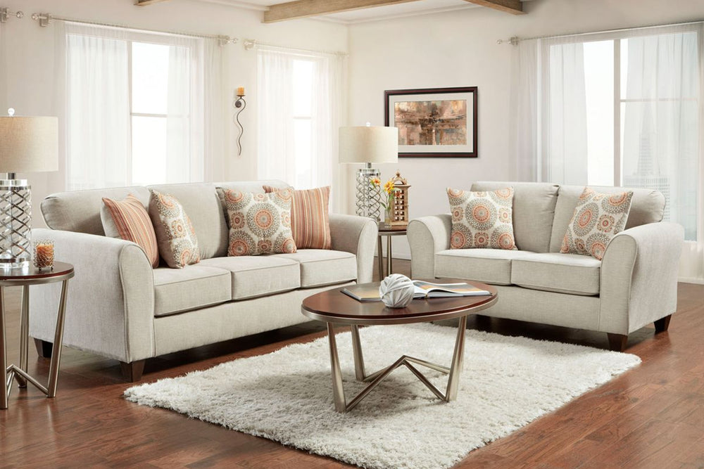 Sala de estar luminosa con sofá y loveseat color beige, cojines decorativos estampados, mesa de centro redonda de madera y aluminio, alfombra blanca y lámparas laterales junto a grandes ventanas con cortinas translúcidas.