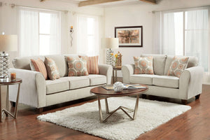 Sala de estar luminosa con sofá y loveseat color beige, cojines decorativos estampados, mesa de centro redonda de madera y aluminio, alfombra blanca y lámparas laterales junto a grandes ventanas con cortinas translúcidas.
