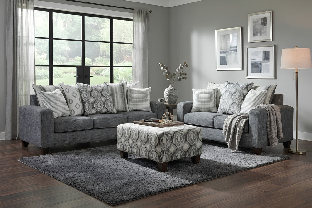 Juego de sala moderno compuesto por sofá y loveseat tapizados en tela gris, con cojines decorativos en patrones geométricos y rayas en tonos gris y blanco, sobre fondo blanco.