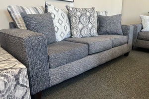 Sofá moderno tapizado en tela gris jaspeada, con cojines decorativos en patrones geométricos y texturas en tonos gris, blanco y negro, exhibido en una tienda de muebles.